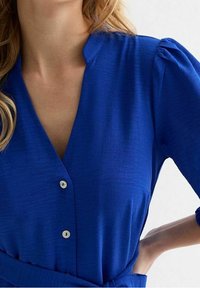 Indumento blu reale con scollatura a V, maniche a sbuffo corte e bottoni visibili sul davanti. Realizzato in un tessuto testurizzato.