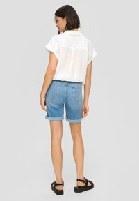 Biała koszula krótki rękaw z kołnierzykiem, zestawiona z niebieskimi dżinsowymi szortami z podwiniętymi mankietami oraz czarnymi sandałami na paski. Neutralne szare tło.