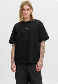Sort bomulds t-shirt med rund hals og korte ærmer, med et lille hvidt logo foran. Modellen har synlige tatoveringer.