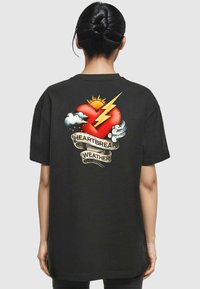 Svart oversized t-skjorte med et grafisk motiv av et rødt hjerte med skyer, lyn og sol, med teksten "HEARTBREAK WEATHER" under.