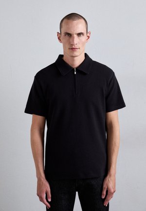 Svart kortärmad polo med dragkedja, tillverkad av strukturerat tyg. Modellen står mot en enkel bakgrund.