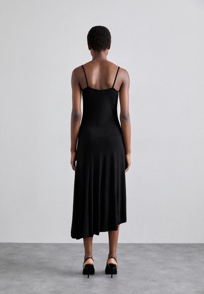 Rochie neagră fără mâneci, cu bretele subțiri, cu o lungime până la genunchi și textură fină. Modelul poartă pantofi cu toc înalt negri. Vedere din spate.
