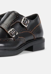 Zwarte leren schoenen met een ronde neus, voorzien van een decoratief perforatiepatroon, twee metalen gespen en een lage blokhak.