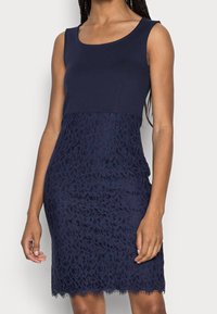 Femme portant une robe sans manches bleu marine avec un haut uni et une jupe en dentelle florale, se tenant devant un fond blanc.