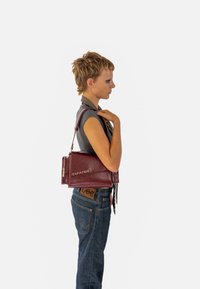 Burgundská kožená kabelka so štruktúrovaným dizajnom, s strieborným zipsom a s názvom značky "EXPATRIÉ" vyrazeným na prednej strane.