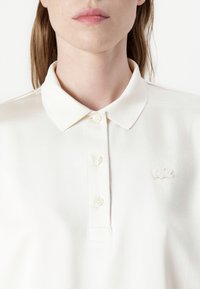 Valge polo särk klassikalise kraega, millel on kolm nuppu ja tikandiga logo vasakul küljel. Sile kangastekstuuri.