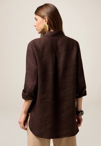 Camisa de lino marrón con mangas enrolladas, cuello y dobladillo curvado. Cuenta con una costura de yugo en la parte posterior y se lleva con pantalones beige y grandes pendientes de oro.