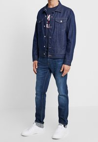 Denim jacket in dark blue with button closure, two chest pockets, and button cuffs. Paired with blue jeans and white sneakers.
