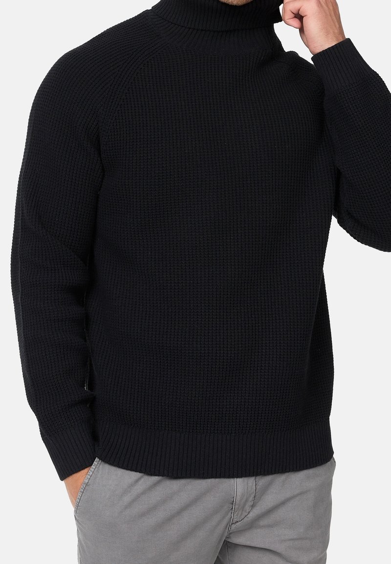 Pull à col roulé noir en maille avec poignets et ourlet côtelés. Le tissu texturé présente un motif de carrés en relief, offrant une apparence chaleureuse.