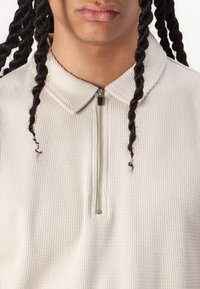 Lys beige, struktureret pullover med krave og kvart lynlås fortil. Har små detaljer på lynlåsen og et ribbet stofmønster.