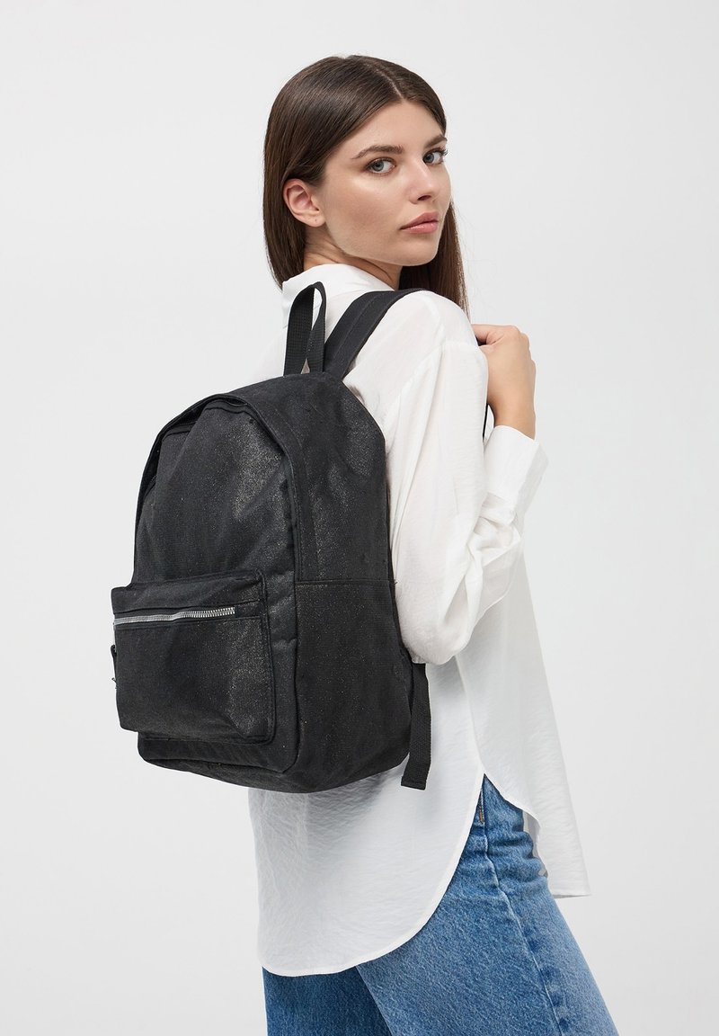 Zaino di tessuto nero con tasca frontale con zip, forma rettangolare, spallacci robusti e finitura texturizzata, indossato su una spalla.