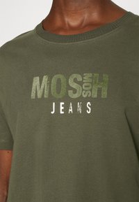 Gros plan sur un t-shirt vert olive avec le logo "MOSH JEANS" imprimé en vert délavé et blanc sur la poitrine.
