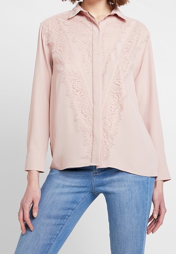 Blouse rose clair à manches longues avec un détail en dentelle florale en forme de V, portée avec un jean bleu clair, modèle posant les mains détendues le long du corps.
