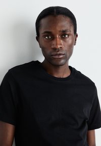 T-shirt en coton noir avec un col rond, des manches courtes et une texture lisse, présenté sur un fond gris clair.
