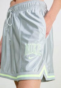 Nike shorts in light gray satin with neon green logo and accents. Features a drawstring waistband and side pockets. Horizontal stripes at hem.