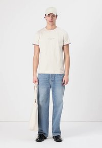 Beige bomull t-skjorte med korte ermer og røde detaljer, lyseblå jeans, svarte sko og en beige caps. Holder en lys tote bag.