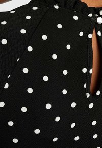 Musta kangas valkoisilla pilkuilla, jossa on avainreikäkiinnitys ja röyhelöillä varustettu kaula-aukko. Pinta on hieman ryppyinen.