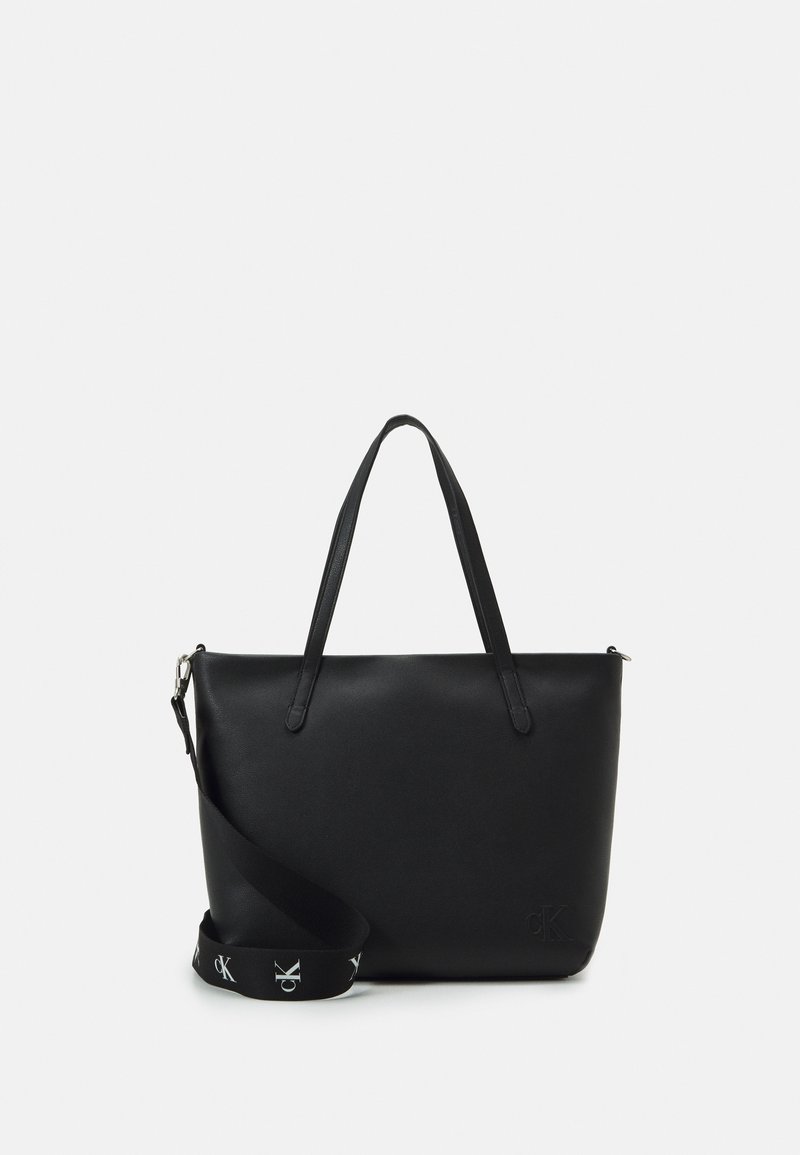 Sac cabas rectangulaire en cuir noir avec deux poignées, bandoulière amovible ornée de logos blancs "cK" et logo "cK" délicatement embossé en bas à droite.