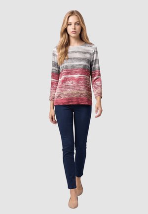 Joven mujer caminando hacia adelante con una camiseta de manga larga a rayas en tonos rojos, grises y beige, pantalones oscuros y zapatos beige sobre un fondo liso.