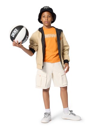 Beige jas met zwarte voering, oranje T-shirt, witte cargobroek met zakken en witte sneakers. Houdt een zwart-witte basketbal vast.