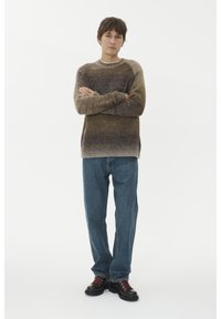 Un jeune homme portant un pull ombré marron, un jean bleu et des bottes sombres se tient les bras croisés devant un fond blanc.