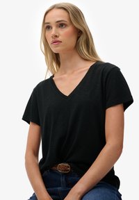 Zwart T-shirt met V-hals en korte mouwen, gemaakt van zachte stof. Voorzien van een subtiel geborduurd logo op de borst. Draagt met een blauwe spijkerbroek en een riem.