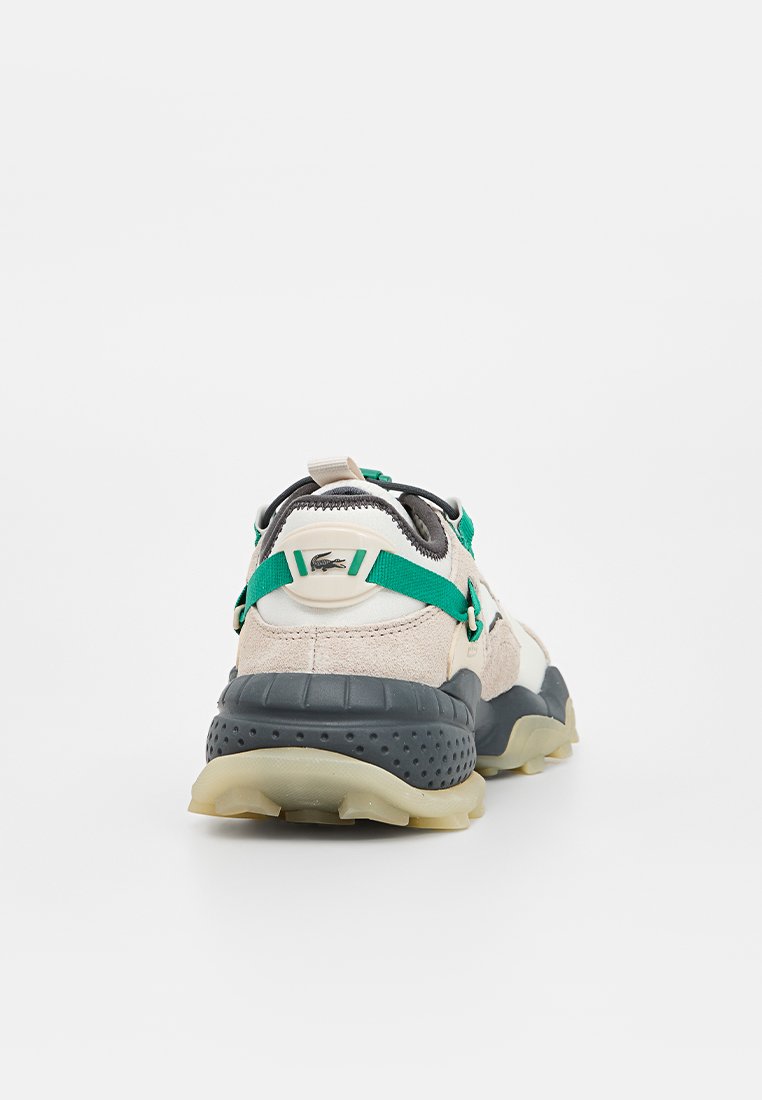 Tył sneakersa z beżową zamszową cholewką, zielonymi akcentami, białą podeszwą środkową i teksturowaną czarną podeszwą. Posiada metkę z logo.