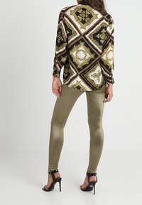 Černá halenka se zlatým vzorem a dlouhými rukávy, doplněná lesklými olivově zelenými legínami a černými sandály na vysokém podpatku s kotníkovými pásky.