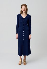 Abito lungo a coste di colore blu navy, con colletto, due tasche frontali e chiusura con bottoni. Dettagli in bottoni color oro e silhouette dritta.