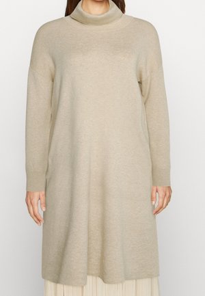 Woman wearing a beige long-sleeve knit sweater dress with a loose turtleneck, standing against a plain background.