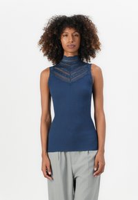 Mulher com cabelo encaracolado, vestindo uma blusa sem mangas de renda azul-marinho e calças cinza clarinho com riscas verticais, em pé contra um fundo branco.