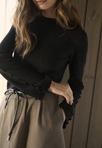 Schwarzer gerippter Strickpullover mit kontrastierenden, geknöpften Bündchen, kombiniert mit hoch taillierten, karierten braunen Hosen mit einem Kordelzugbund.