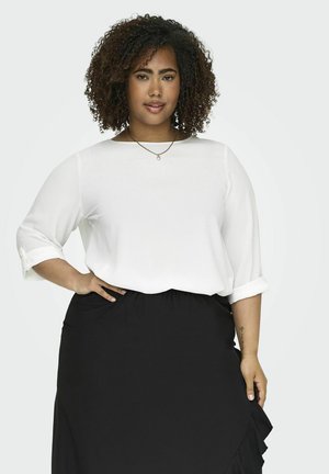 Blouse blanche à manches trois-quarts, avec des poignets repliés. Associée à une jupe noire avec un ourlet à volants. Texture de tissu lisse.