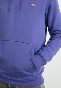 Haut à capuche en polaire violet avec une poche avant, des poignets côtelés et un ourlet côtelé. Présente un patch logo sur la poitrine. Texture douce, design décontracté.