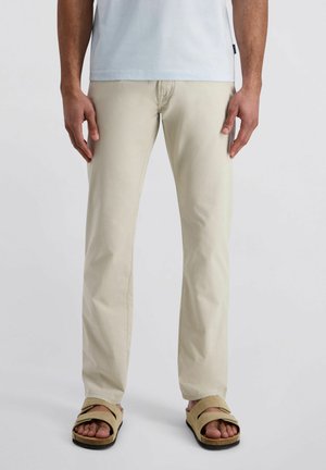 Man draagt beige recht vallende broek, lichtgrijs t-shirt en beige sandalen, staand tegen een effen witte achtergrond.