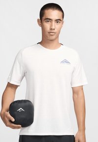 Vit sportskjorta med korta ärmar som har ett strukturerat mönster och en logotyp. Hållen i handen är en kompakt, svart dragsko-påse.