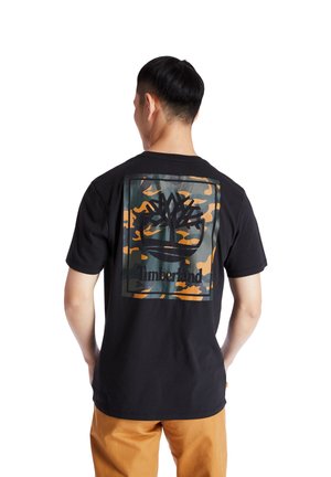 Mann trägt ein schwarzes Timberland T-Shirt mit großem Tarnmuster-Logo auf dem Rücken, kombiniert mit beigen Hosen, stehend vor weißem Hintergrund.