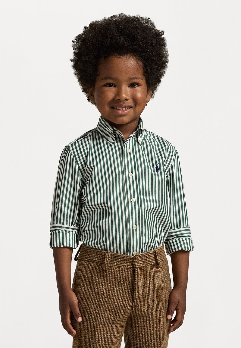 Πράσινο και άσπρο ριγέ πουκάμισο με γυρισμένα μανίκια, συνδυασμένο με υφασμάτινες καφέ καρό παντελόνες. Παιδί με σγουρά μαλλιά χαμογελά στην κάμερα.