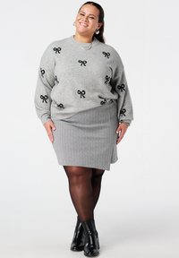 Femme souriante portant un pull gris avec des nœuds noirs, une jupe grise, des collants noirs et des bottines noires, debout devant un fond blanc.