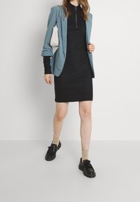 Svart ribbad klänning i kombination med en ljusblå kavaj, svarta skor med fransar och beige strumpor. Outfiten har en dragkedja vid halsen.