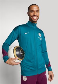 Teal zip-up takki vaaleanpunaisilla yksityiskohdilla ja valkoisella Nike-logolla, yhdistettynä viininpunaisiin housuihin. Pitää kädessään valkoista ja mustaa jalkapalloa kultaisin yksityiskohdin.