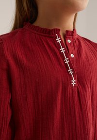 Top in tessuto rosso testurizzato con colletto arricciato, patta con bottoni e cuciture decorative bianche lungo il centro.