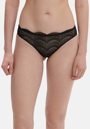 Kobieta ubrana w czarne koronkowe figi z falowanymi krawędziami, stojąca na białym, jednolitym tle.