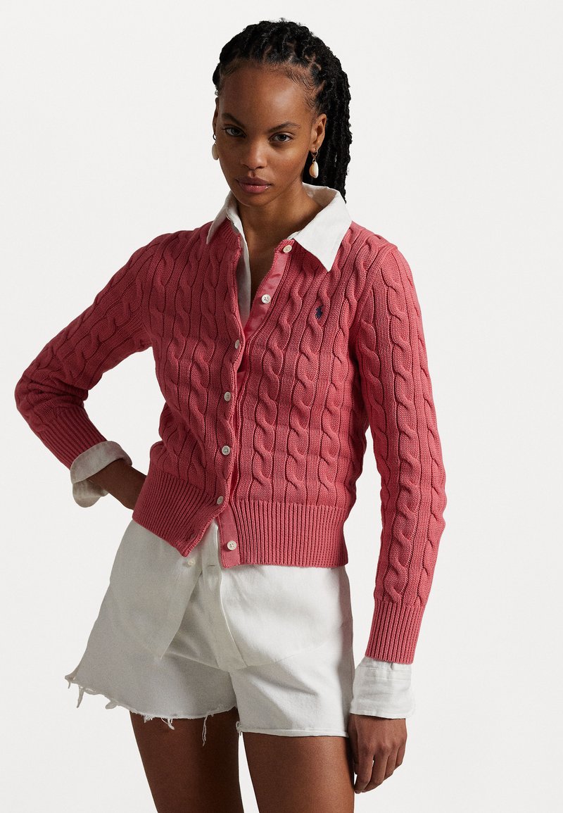 Rosa Kabelstrick-Cardigan mit geripptem Saum, darunter ein kragenweißes Hemd. Mit Knöpfen vorne und einem bestickten Logo-Detail.