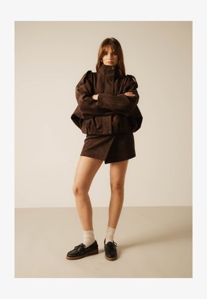 Kvinne står med armene i kors iført en oversized mørk brun jakke, matchende skjørt, hvite sokker og svarte snøresko mot en ensfarget bakgrunn.