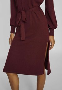 Burgundy midiklänning med långa ärmar, bälte vid midjan och sidoslits. Tyget verkar mjukt med en lätt textur som förbättrar den kroppsnära designen.