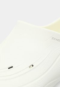 Baltas slip-on apavas ar gludu, teksturētu virsmu, izliekts formas, ventilācijas caurumi un neuzbāzīgas viļņu raksti augšējā materiālā.