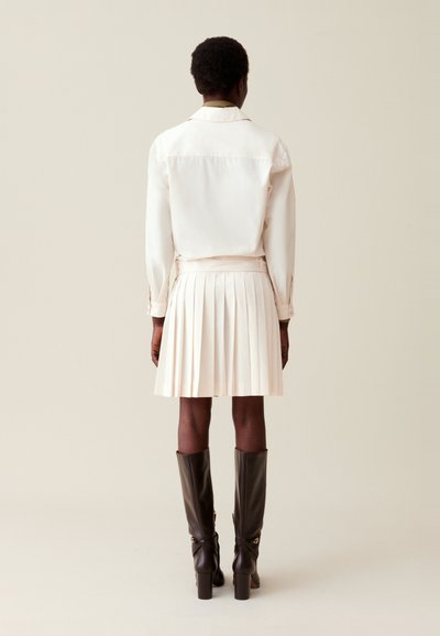 Jupe plissée blanche et chemise assortie avec un col, assorties à des bottes marron montantes. Le tissu semble lisse avec des détails soignés.