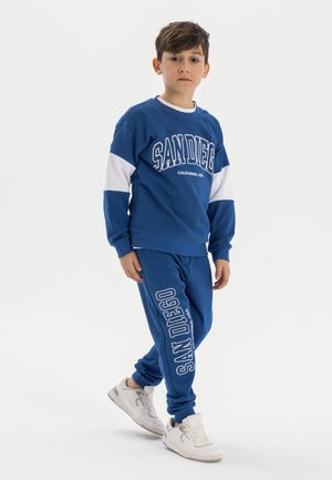 Blue cotton sweatshirt with white accents featuring "SAN DIEGO" print. Matching blue joggers with logo and white detailing. White sneakers.