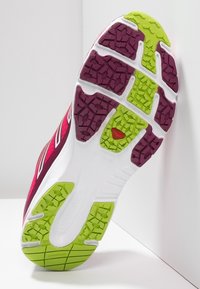 Löparsko med gröna och lila gummitärningar på vit mellansula, som visar ett texturerat greppmönster och röd logodetalj.